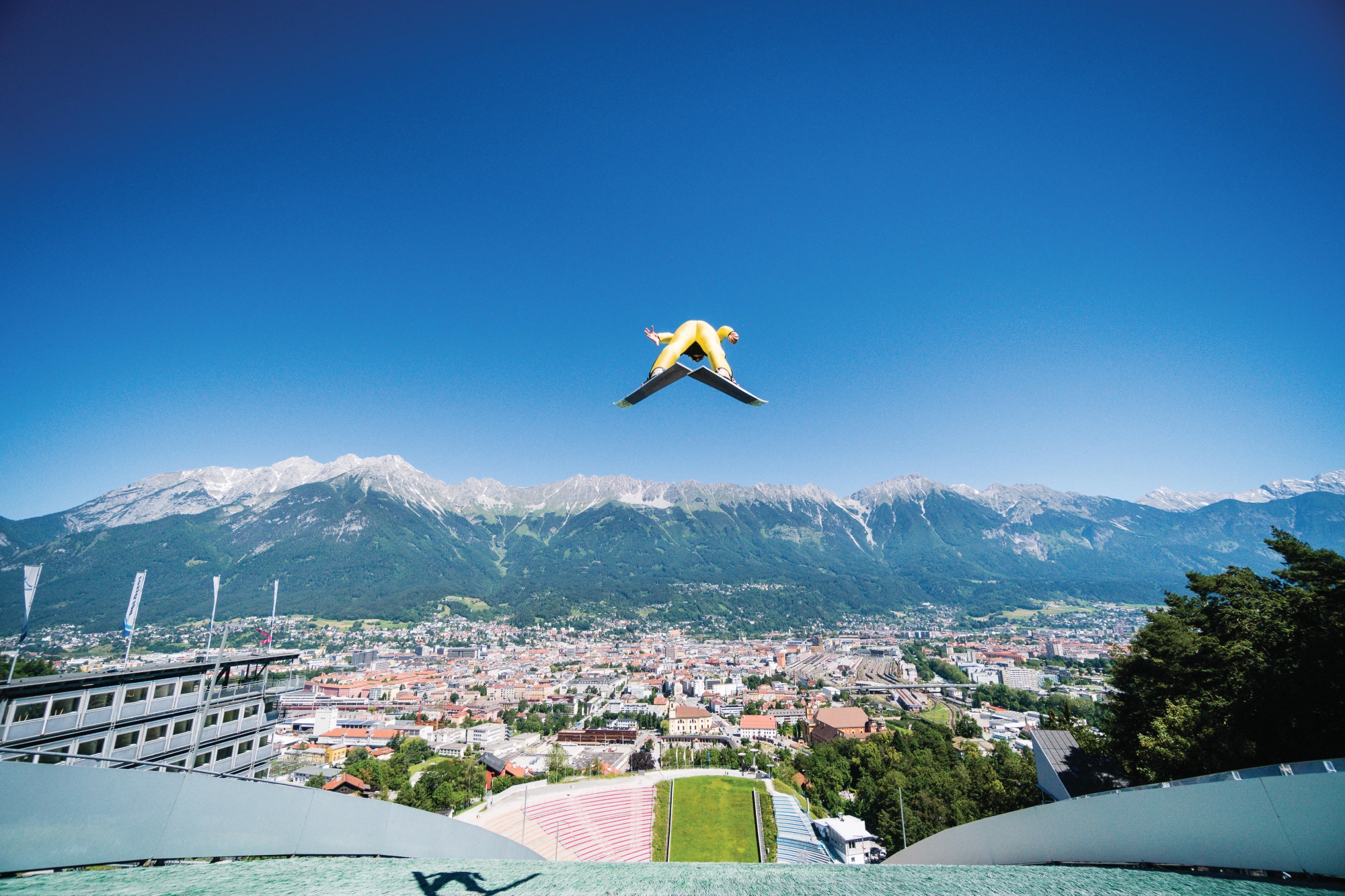Saltatori con gli sci in aria con una vista panoramica sulle Alpi e un cielo azzurro sullo sfondo.